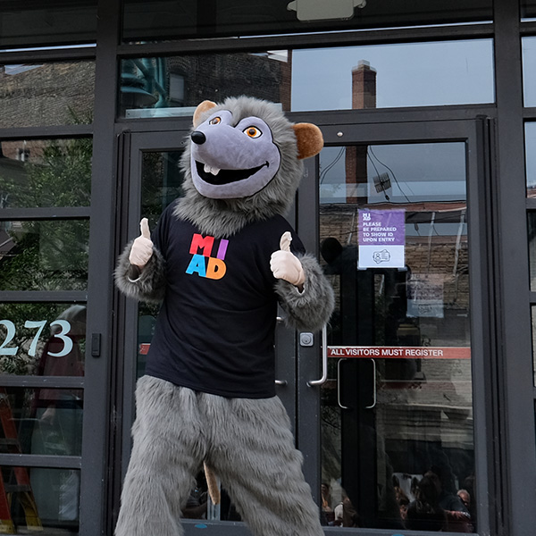 MIAD's River Rat stands in front of the building giving a thumbs up