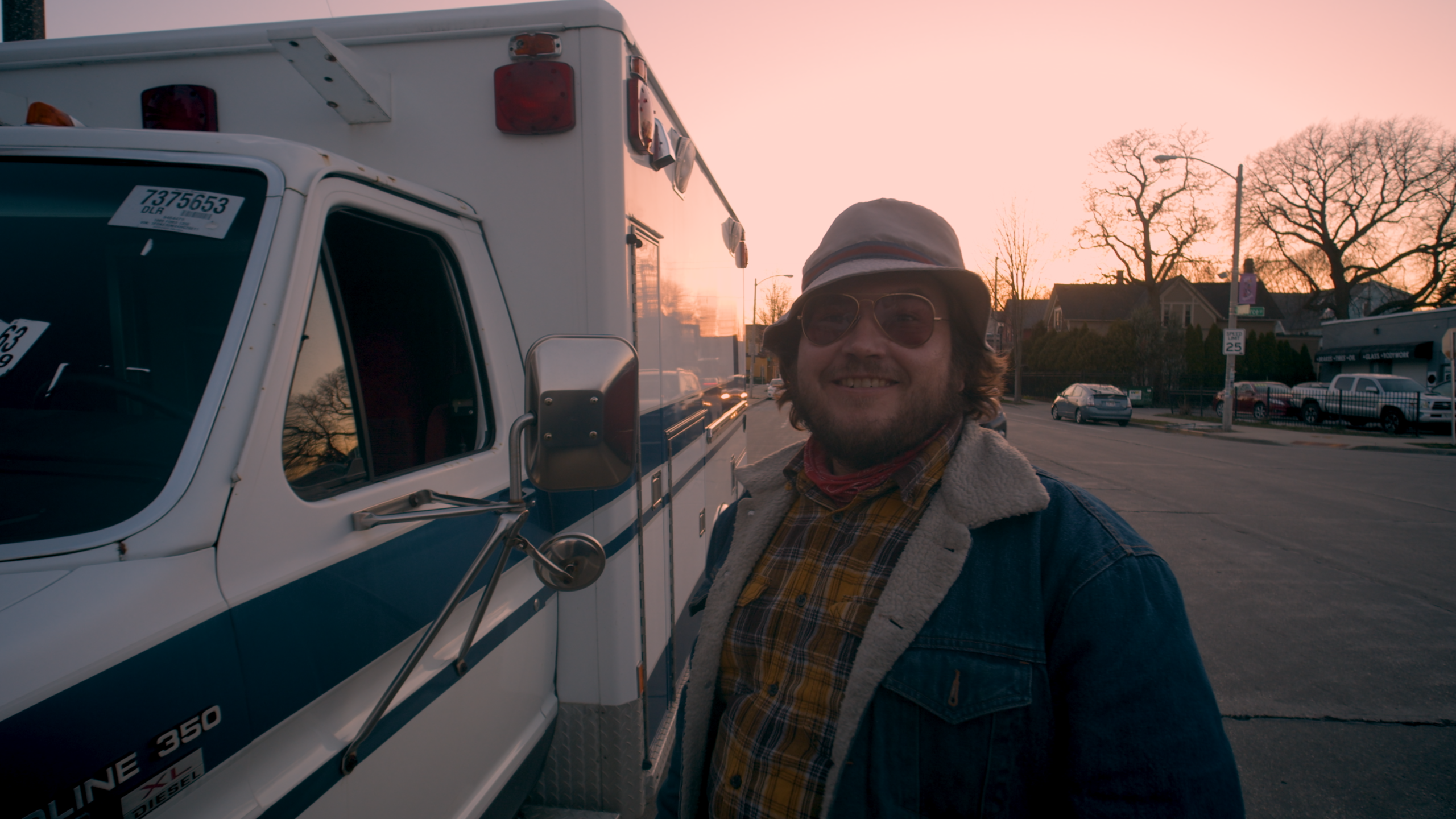 A person wearing a bucket hat, glasses, and a denim jacket stands beside a white utility truck parked on a residential street at sunset. Trees, houses, and parked vehicles are visible in the background.