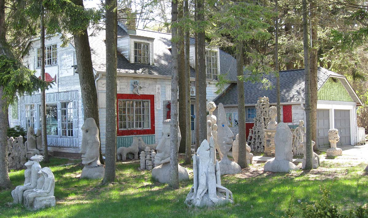 Exterior view of a house surrounded by trees and numerous outdoor sculptures. Stone and concrete artworks of varying sizes are arranged throughout the yard, creating a sculpture garden around the residence.